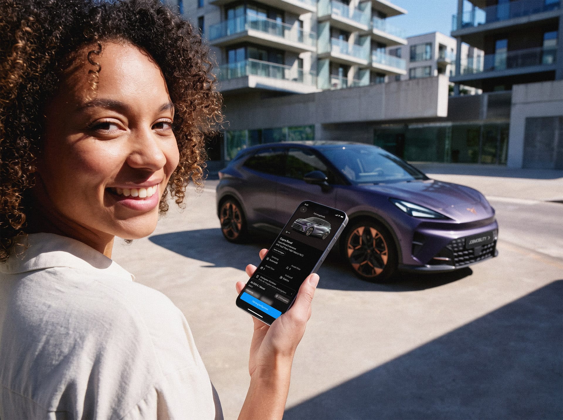 Eine Frau zeigt ein Smartphone mit einer JobMobility-App-Anzeige für einen lila CUPRA Raval. Im Hintergrund steht derselbe CUPRA Raval mit dem Kennzeichen "JOBMOBILITY" vor städtischen Wohngebäuden.