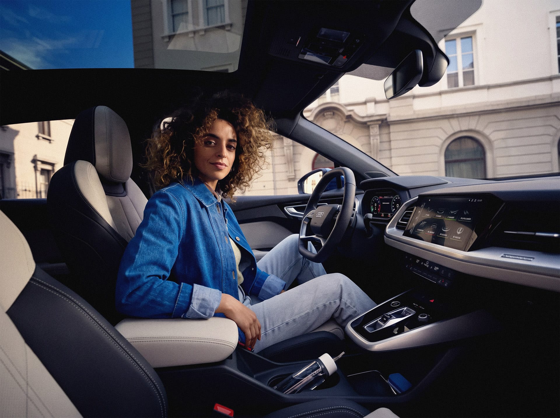Frau in Jeansjacke sitzt lässig im hochmodernen Cockpit eines Audi mit großem Infotainment-Display und Glasdach. Die Atmosphäre vermittelt den Komfort und die Freiheit der JobMobility App.